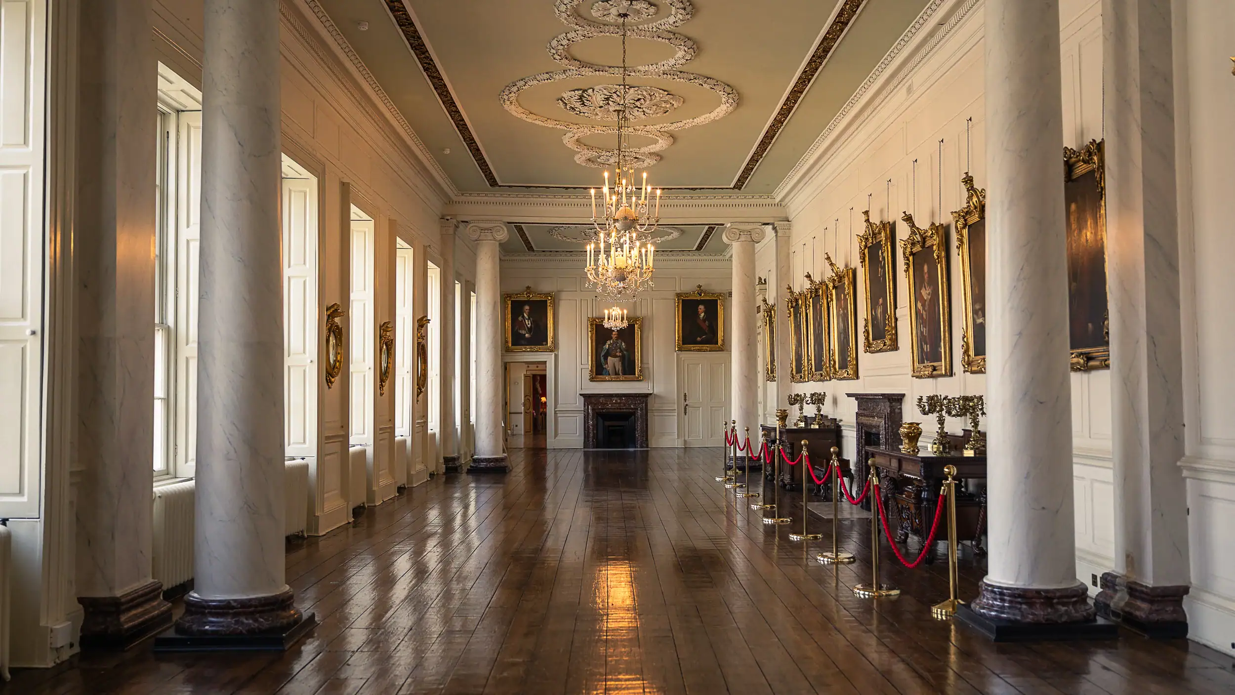 Inside a hallway at Dublin Castle with tall columns, antique paintings, and dark wooden floors reflecting the natural light and chandeliers. A quiet, historic part of the building full of old-world character.