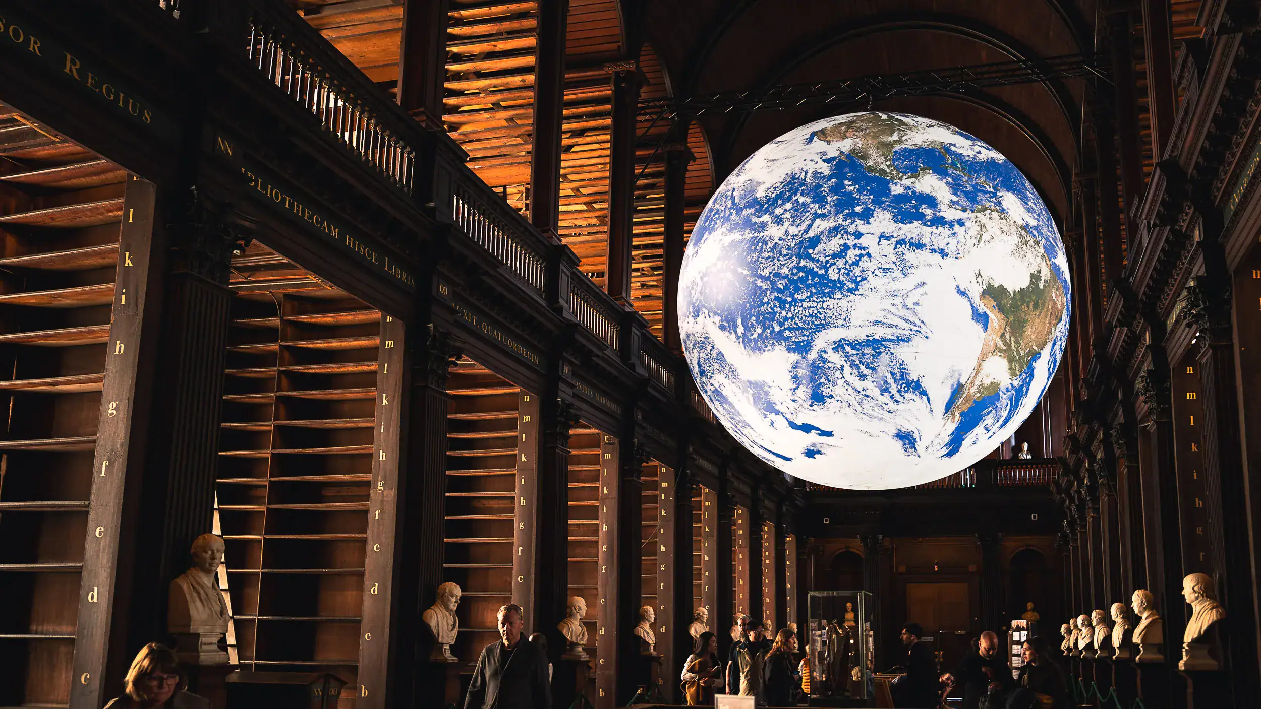 Large illuminated globe art installation (Luke Jerram’s Gaia) suspended inside the Long Room at Trinity College Library in Dublin. The iconic arched space is dimly lit and peaceful, with empty bookshelves as part of a conservation project.