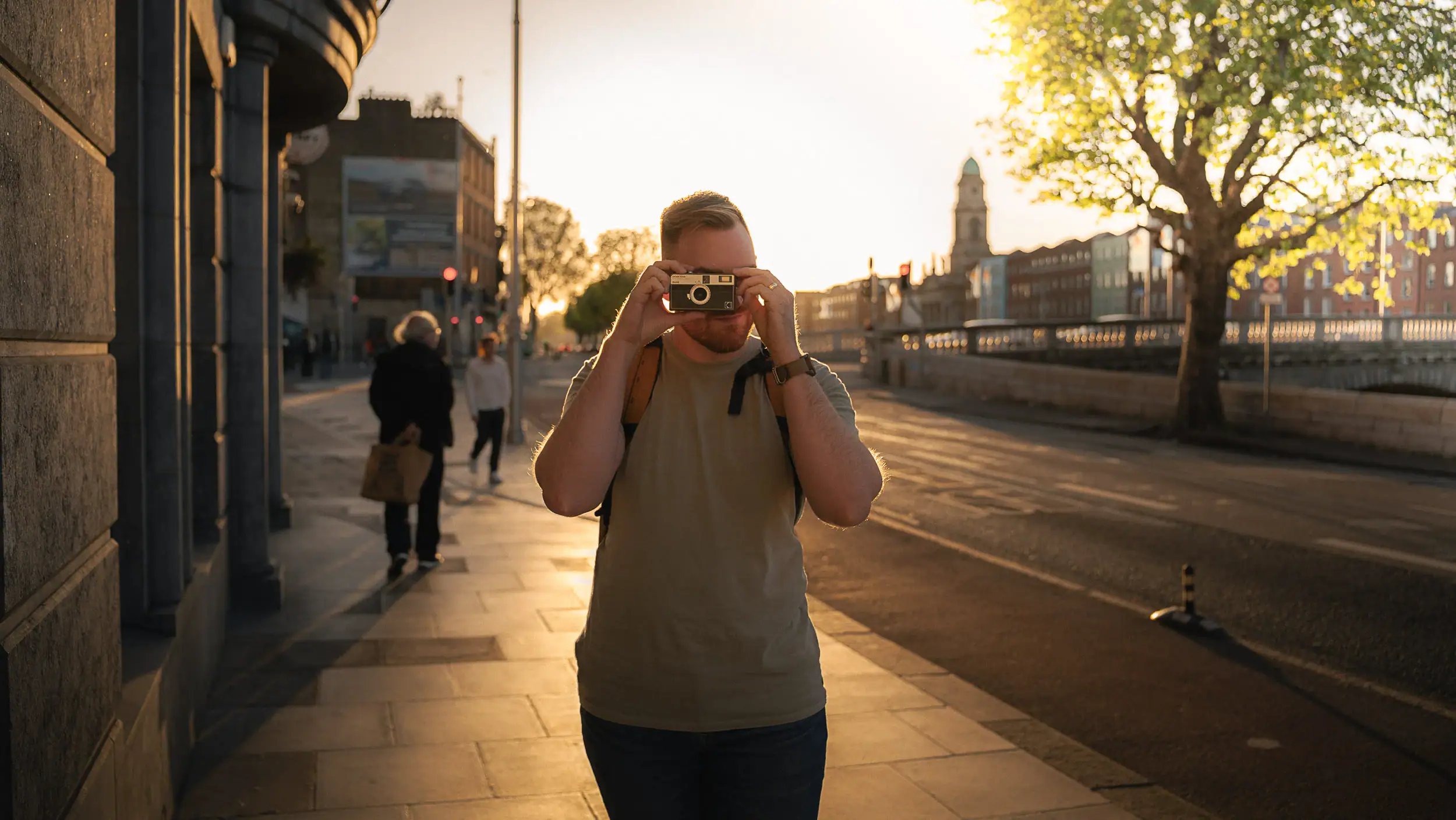 Standing by the river at sunset in Dublin, holding up my film camera with the city’s historic buildings behind me — a real moment from my first time in the city.