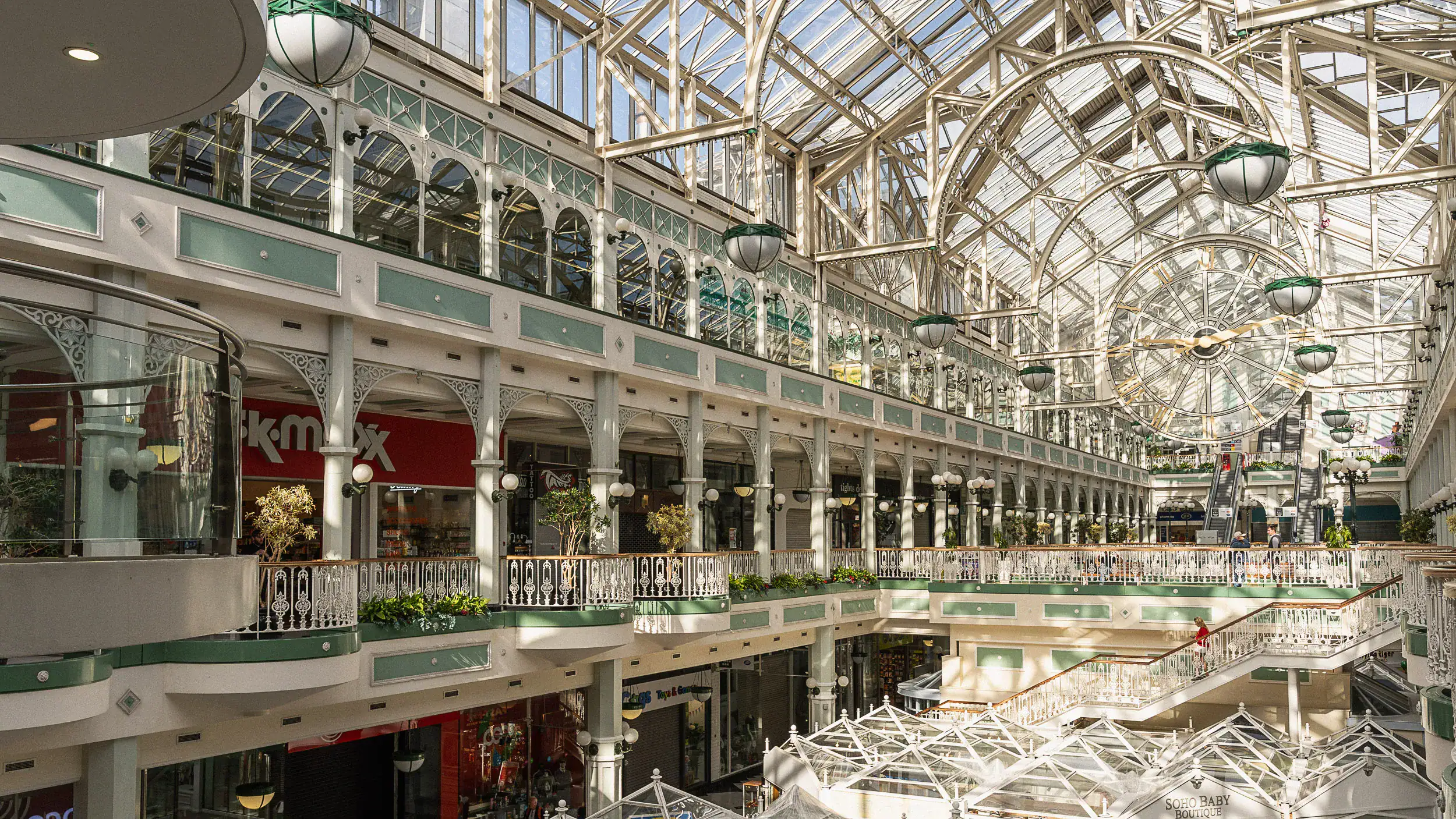 Interior of St. Stephen’s Green Shopping Centre in Dublin. Morning light pours through the glass atrium, reflecting off elegant ironwork and windows. A few people sit quietly on benches beneath the soaring ceiling, soaking in the calm atmosphere of the nearly empty mall.