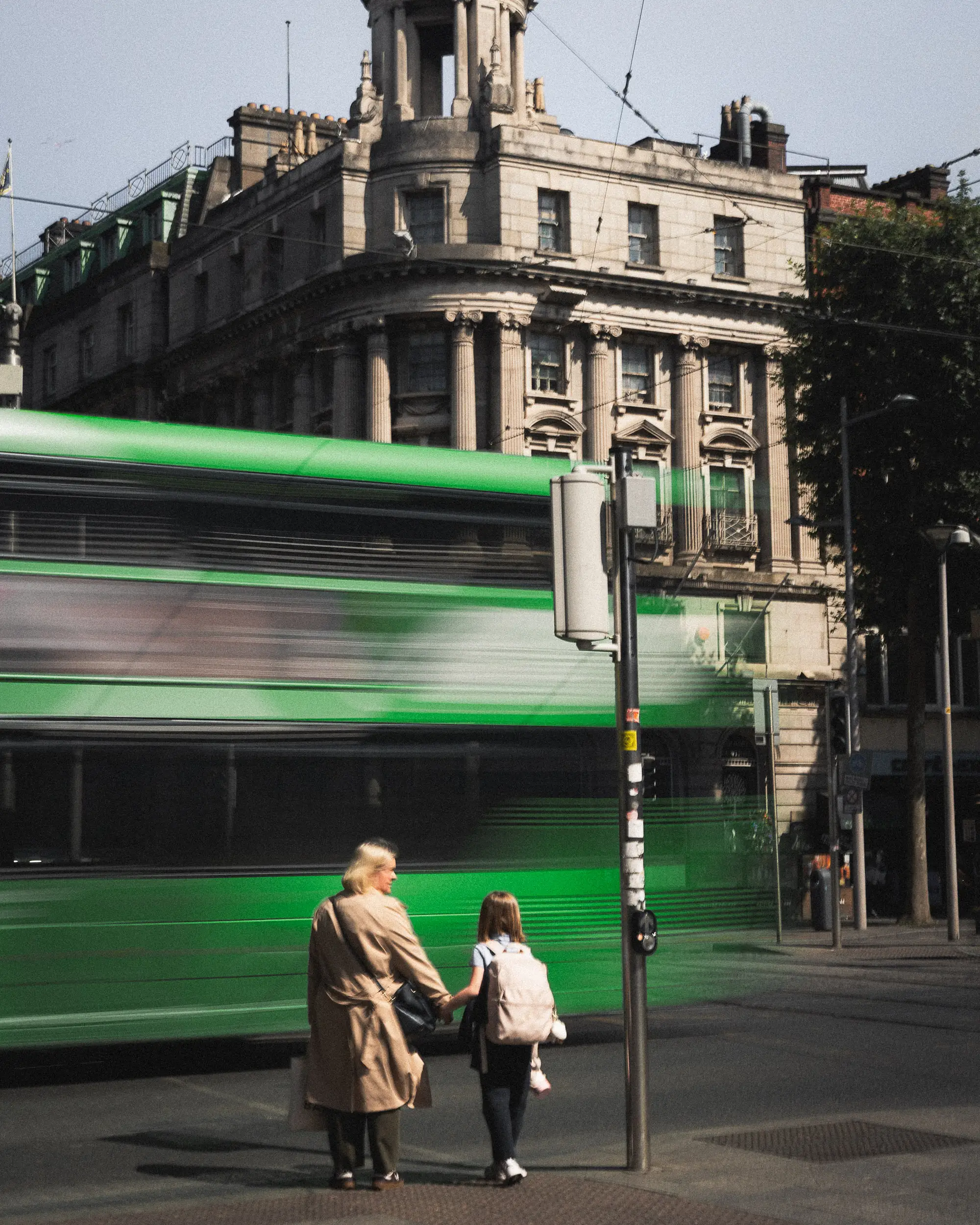 Mother and young daughter holding hands while waiting to cross a busy Dublin street, with a green double-decker bus passing by, capturing the city’s lively pace and family moments.
