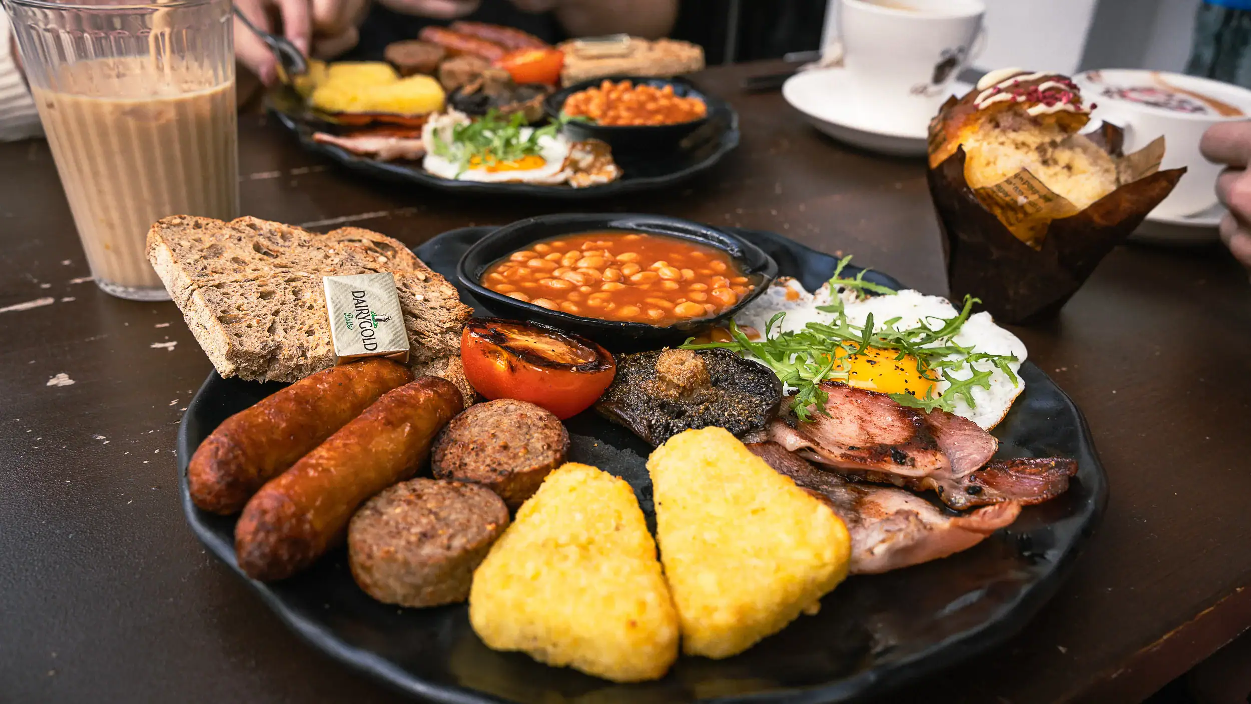 Plate of a traditional full Irish breakfast featuring black and white pudding, grilled tomato, sausage, Guinness bread, baked beans, hashbrowns, and a sunny-side-up egg garnished with pepper.
