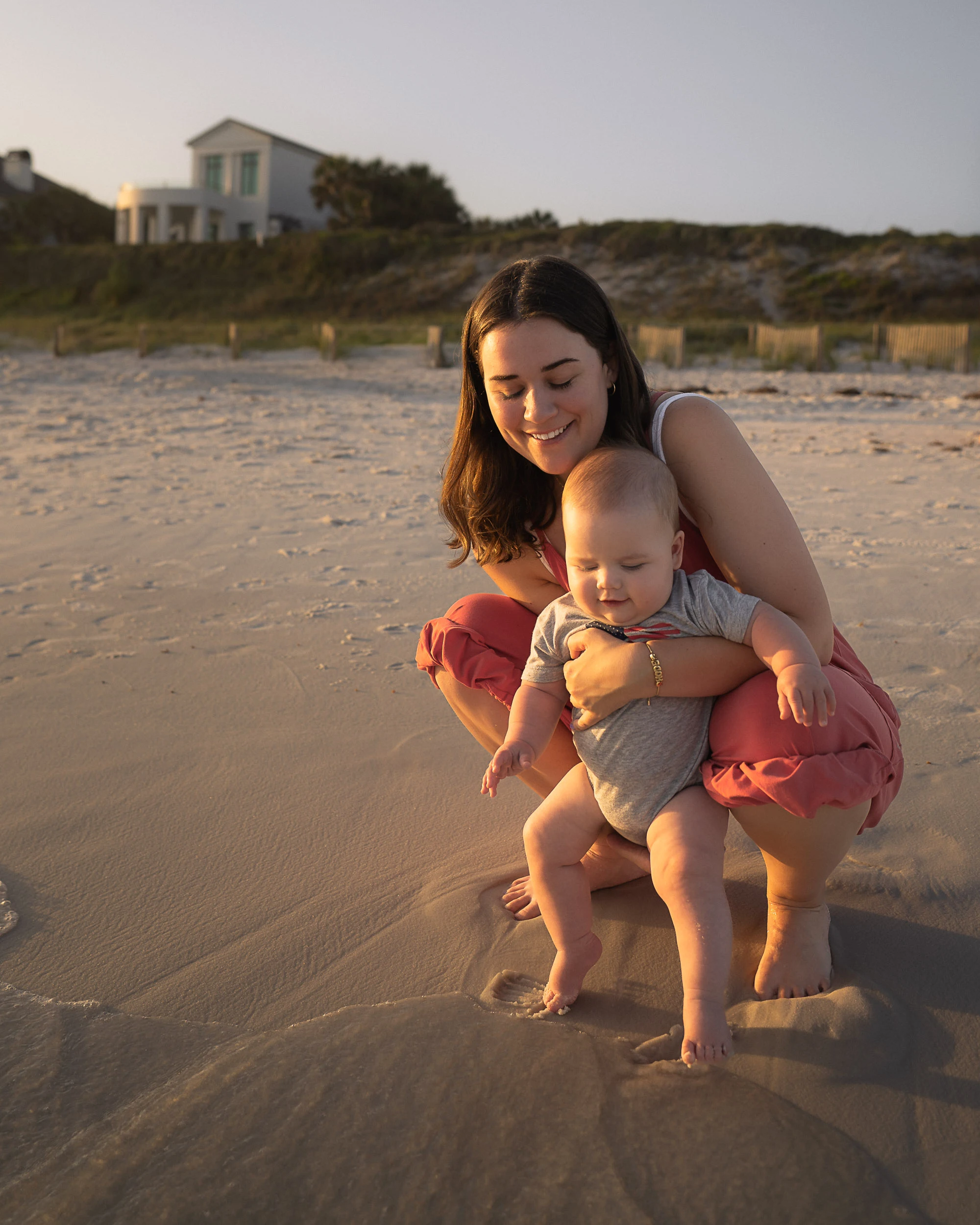 Mother holding smiling baby dipping feet in the water during a calm beach sunset.