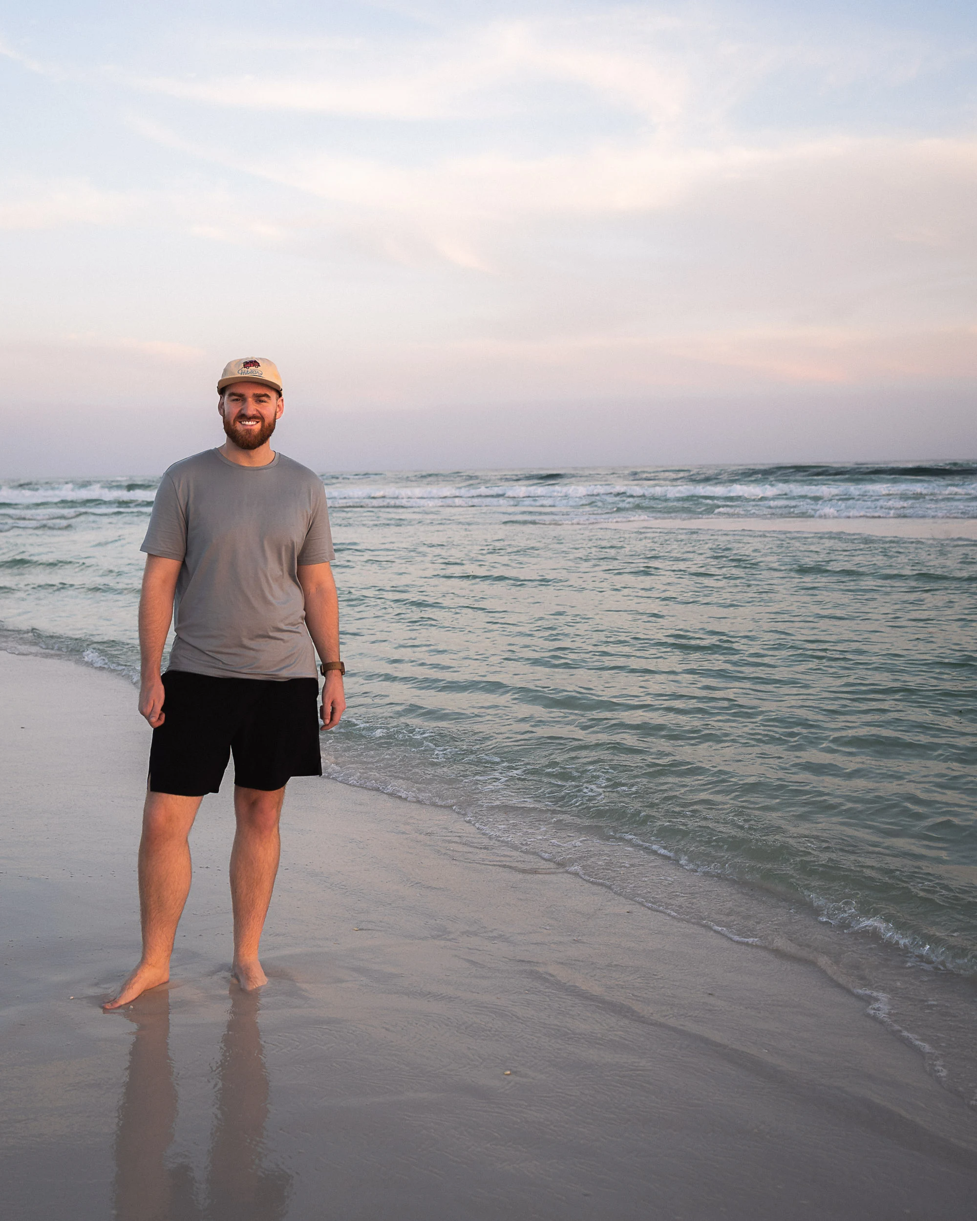 Smiling man standing by emerald waters with a pastel-colored sunset sky in the background.