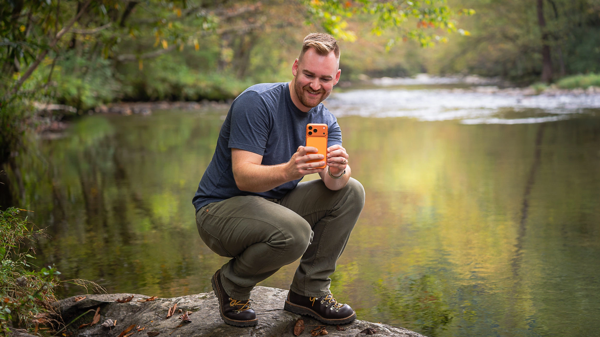 Photographer using iPhone 17 Pro Max to take photos in the Smoky Mountains, squatting on a rock near a calm stream with a smile.