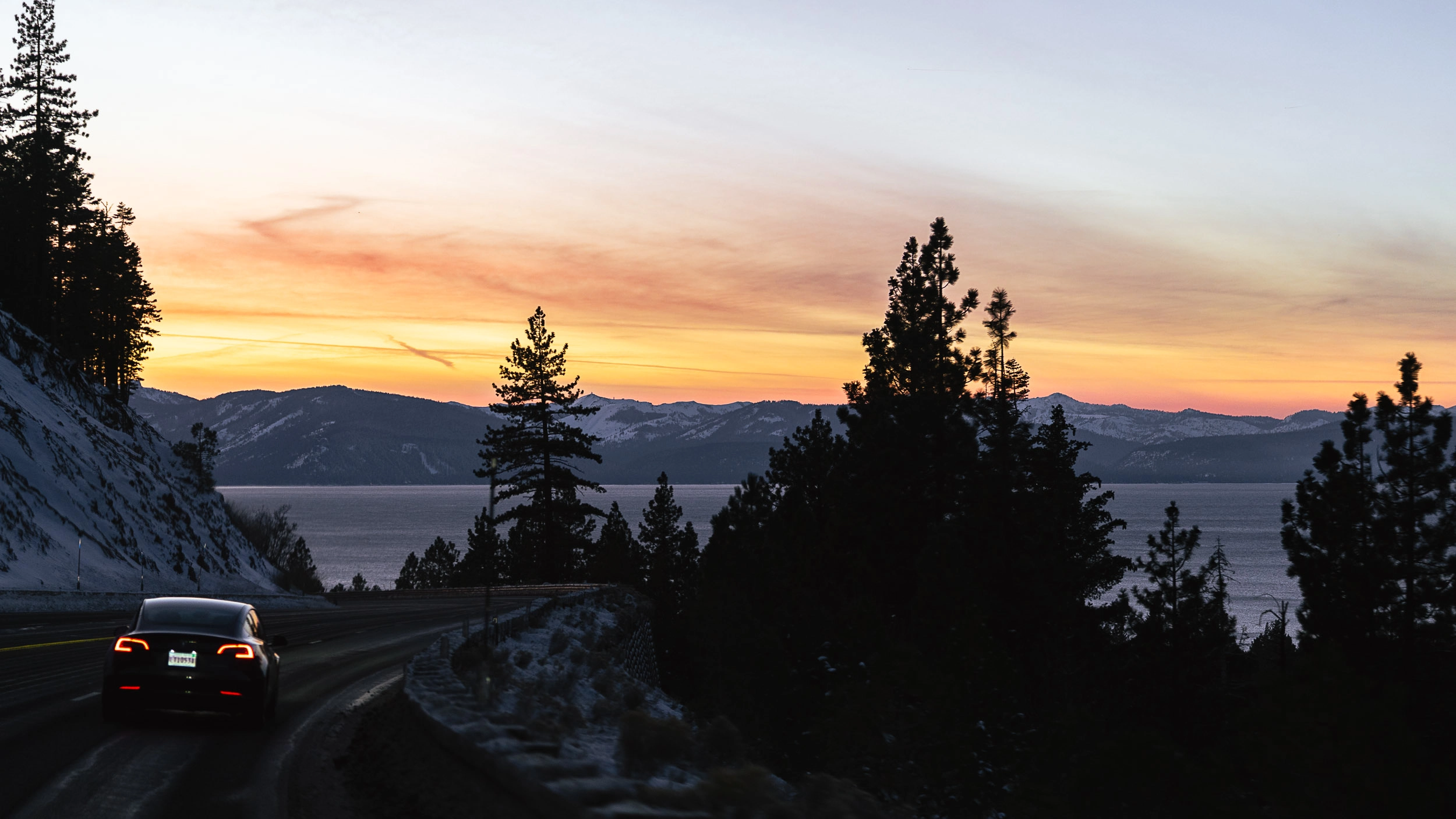 Car driving along a winding Lake Tahoe road at sunset with silhouetted trees and a glowing sky in the background.