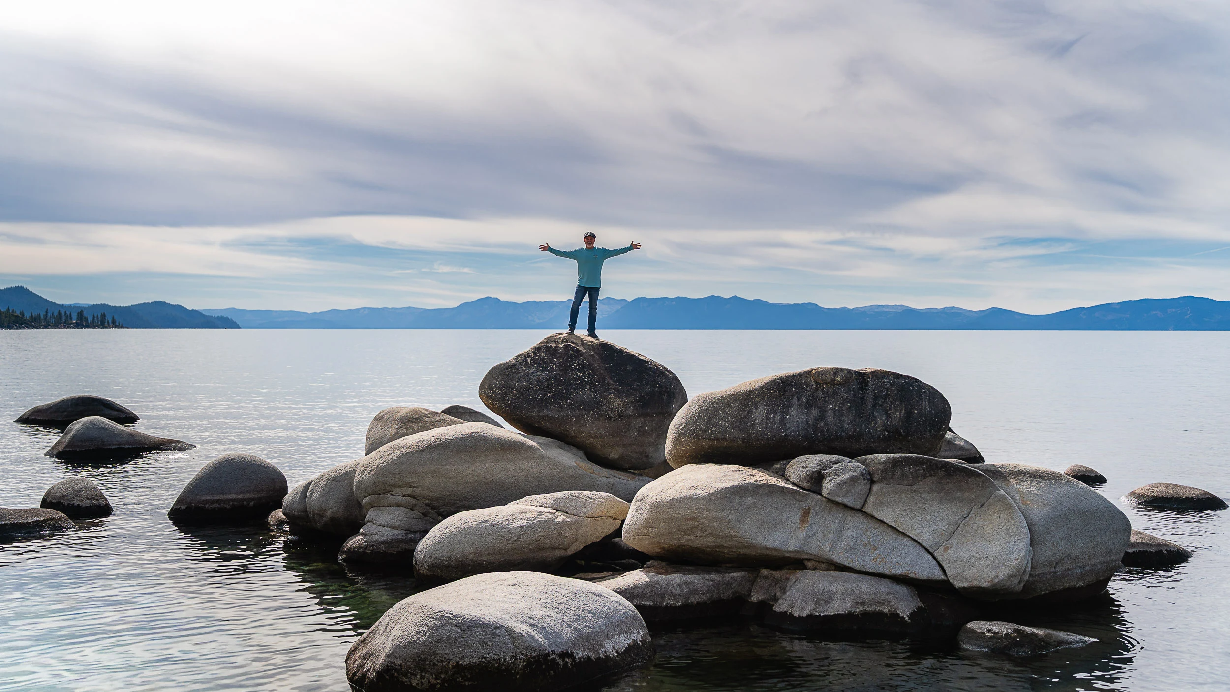 Person standing on Lake Tahoe’s rounded shoreline boulders facing the camera with snow-covered mountains and the lake in the background.