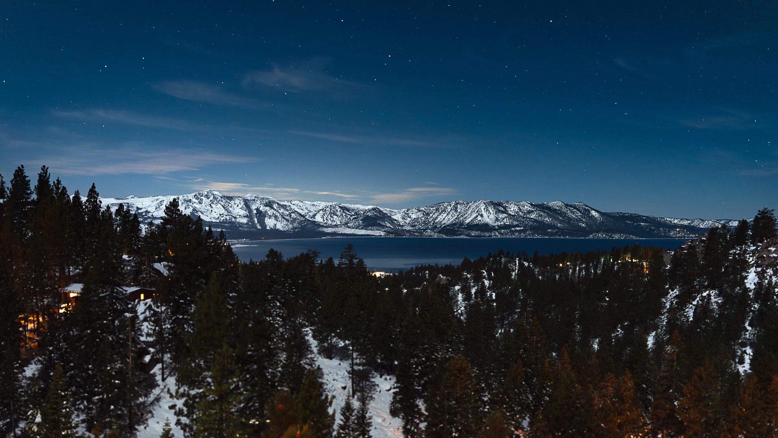 Night view overlooking Lake Tahoe with snowy mountains, a forest in the foreground, and cabin lights glowing around the lake beneath a star-filled sky.